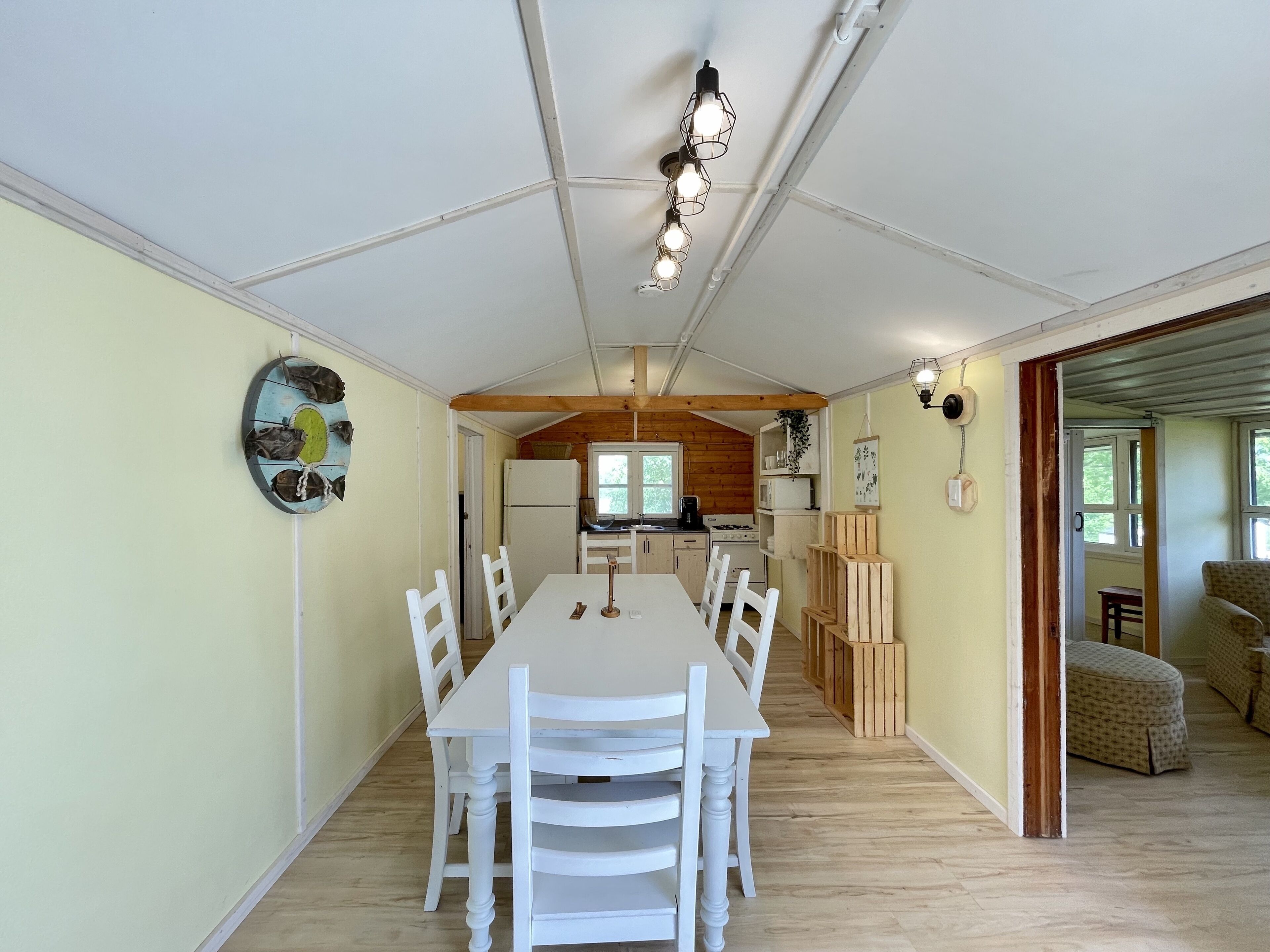 Een eethoek met een lange tafel en witte stoelen, een keuken met houten kasten, en uitzicht door een open deur.
