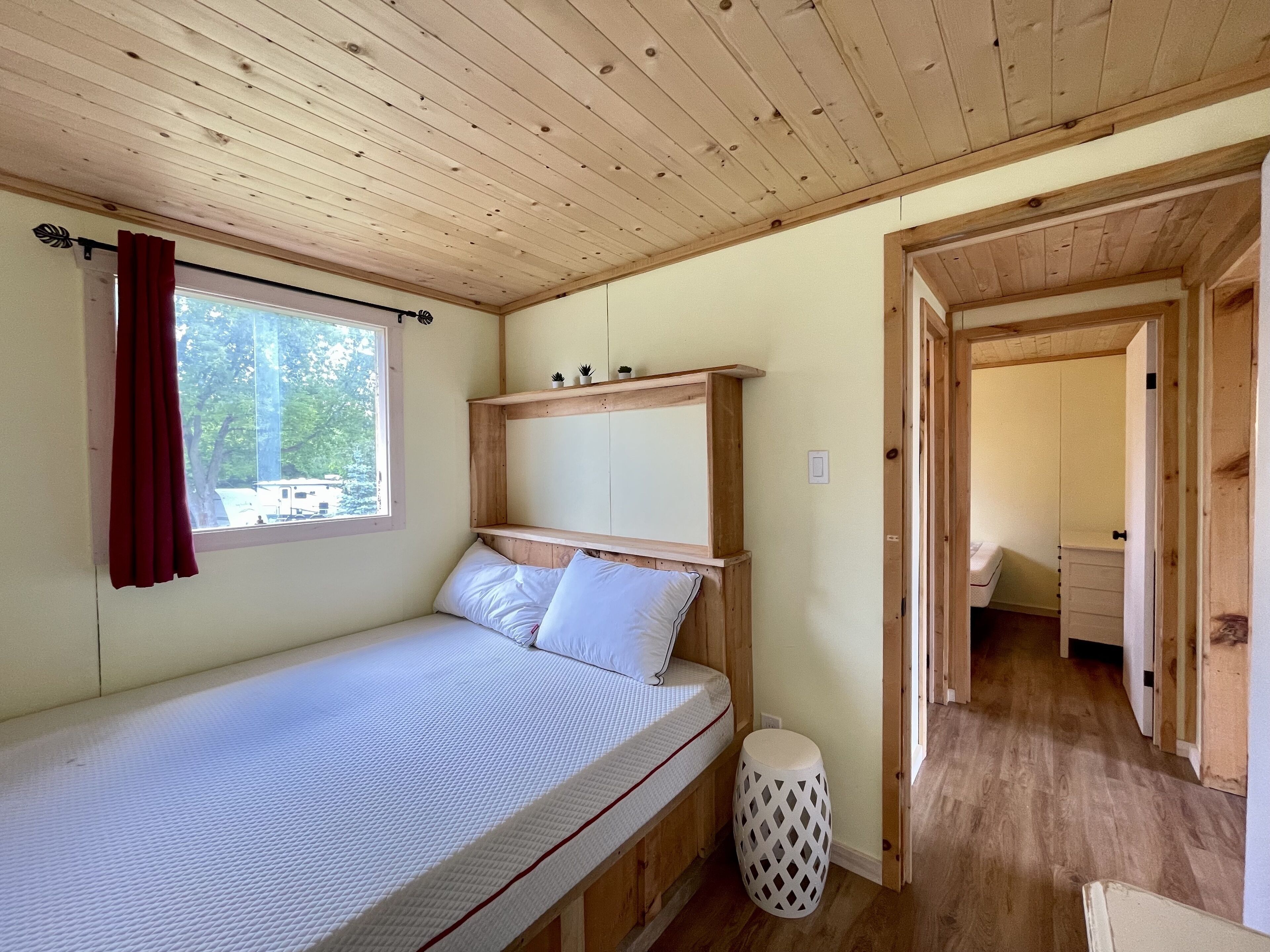 Ein Zimmer mit einem Einzelbett, Holzbalkendecke, einem Fenster mit roten Vorhängen, einem kleinen Nachttisch und einer Tür zu einem anderen Raum.