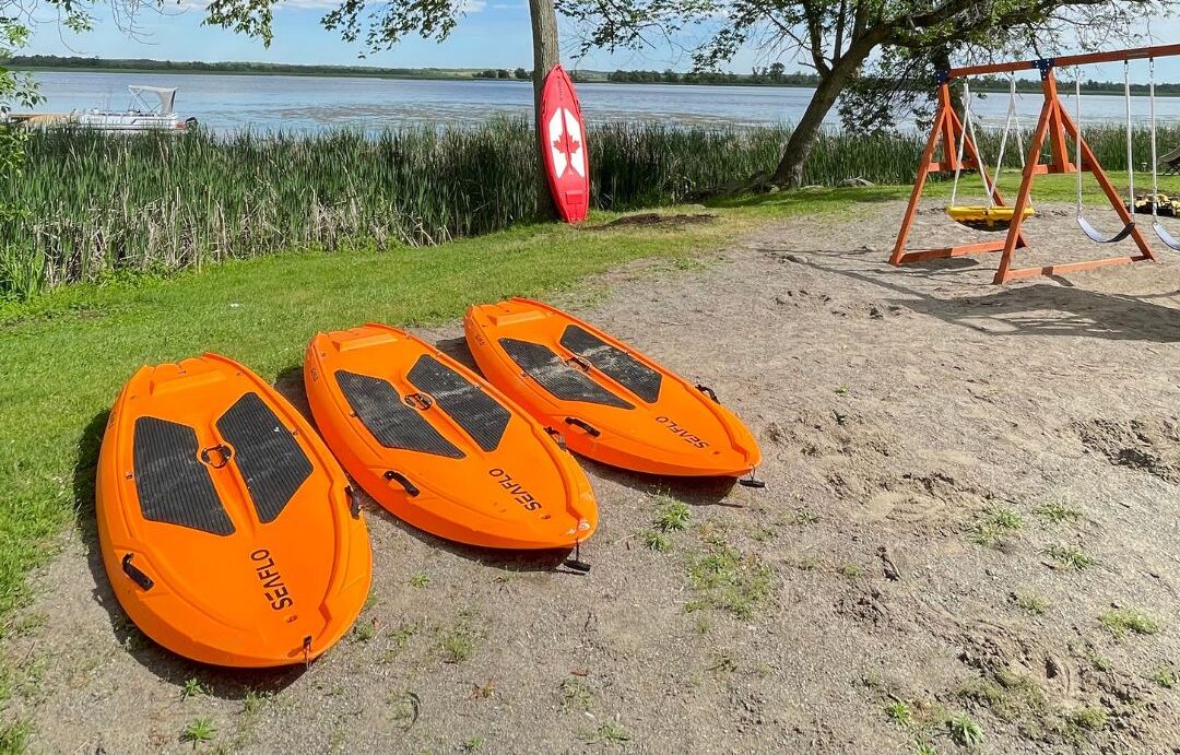 Three orange watercrafts labeled 'SEnFLO' on a sandy shore with a lake and a swing set in the background.