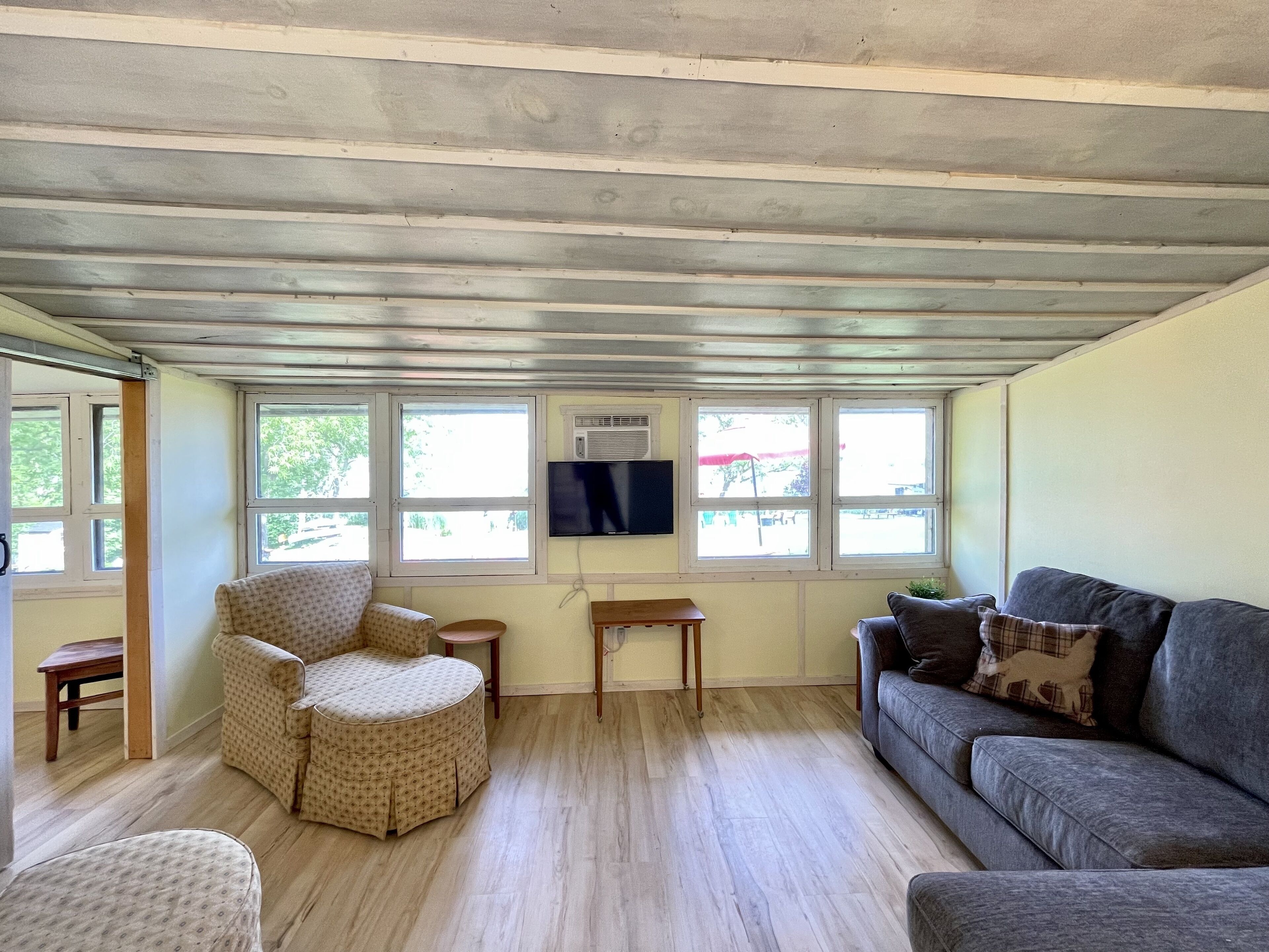 Une pièce avec un plafond en bois, un canapé gris, un fauteuil à motifs, une petite table et un téléviseur fixé au mur.