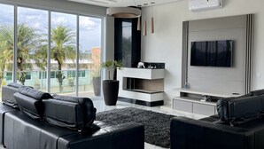 Un salon moderne doté d’un grand canapé d’angle, d’un téléviseur à écran plat fixé au mur et d’une grande fenêtre offrant une vue sur des palmiers et des immeubles.