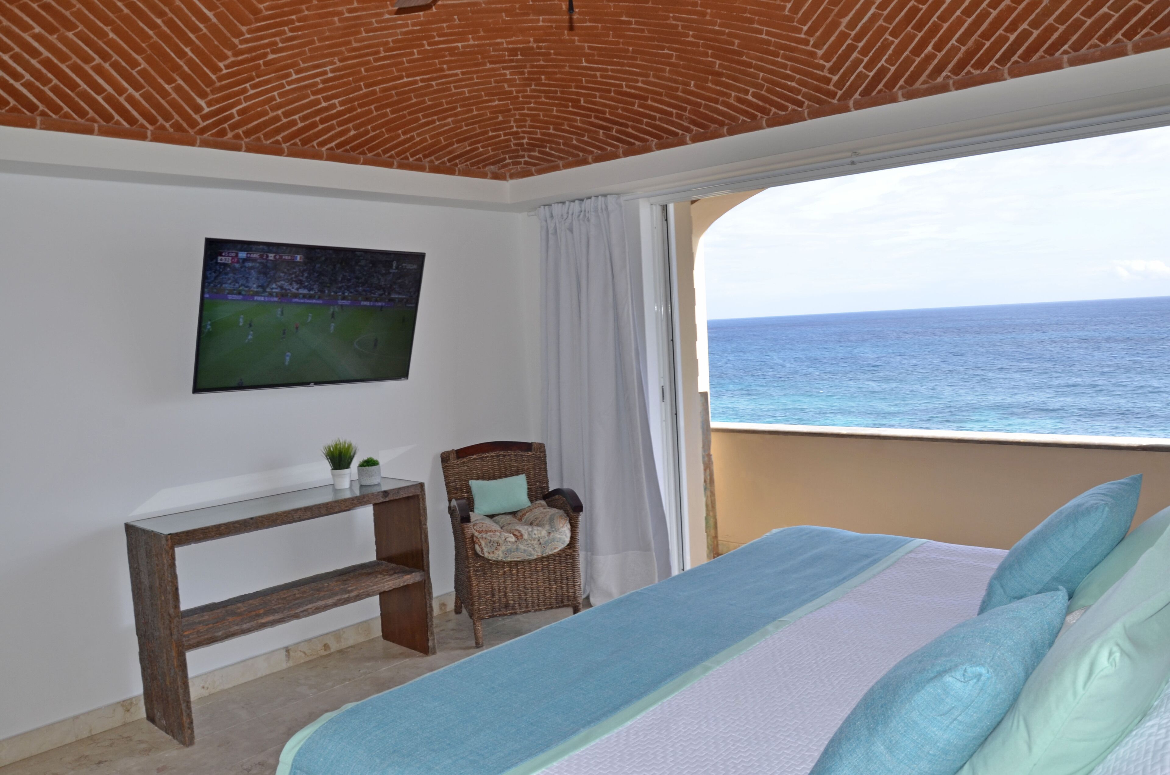 A bedroom with a bed, a TV on the wall, a chair, and a view of the sea.