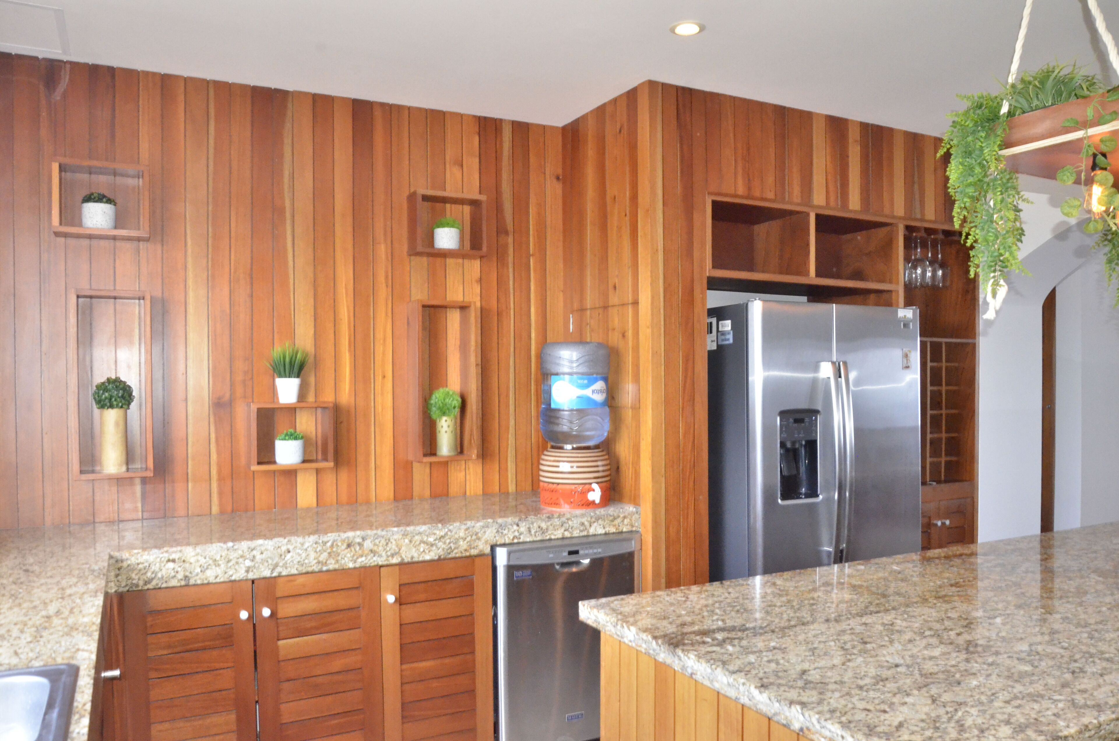 A kitchen with wooden paneling, granite countertops, a stainless steel refrigerator, a dishwasher, and potted plants.