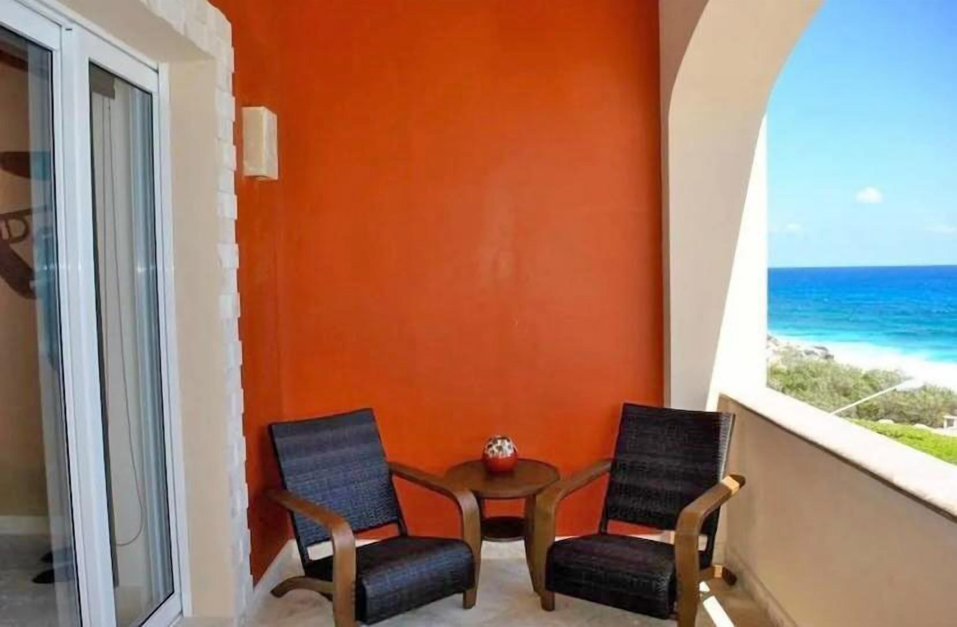 A balcony with two chairs and a small table, overlooking a beach with clear blue water.