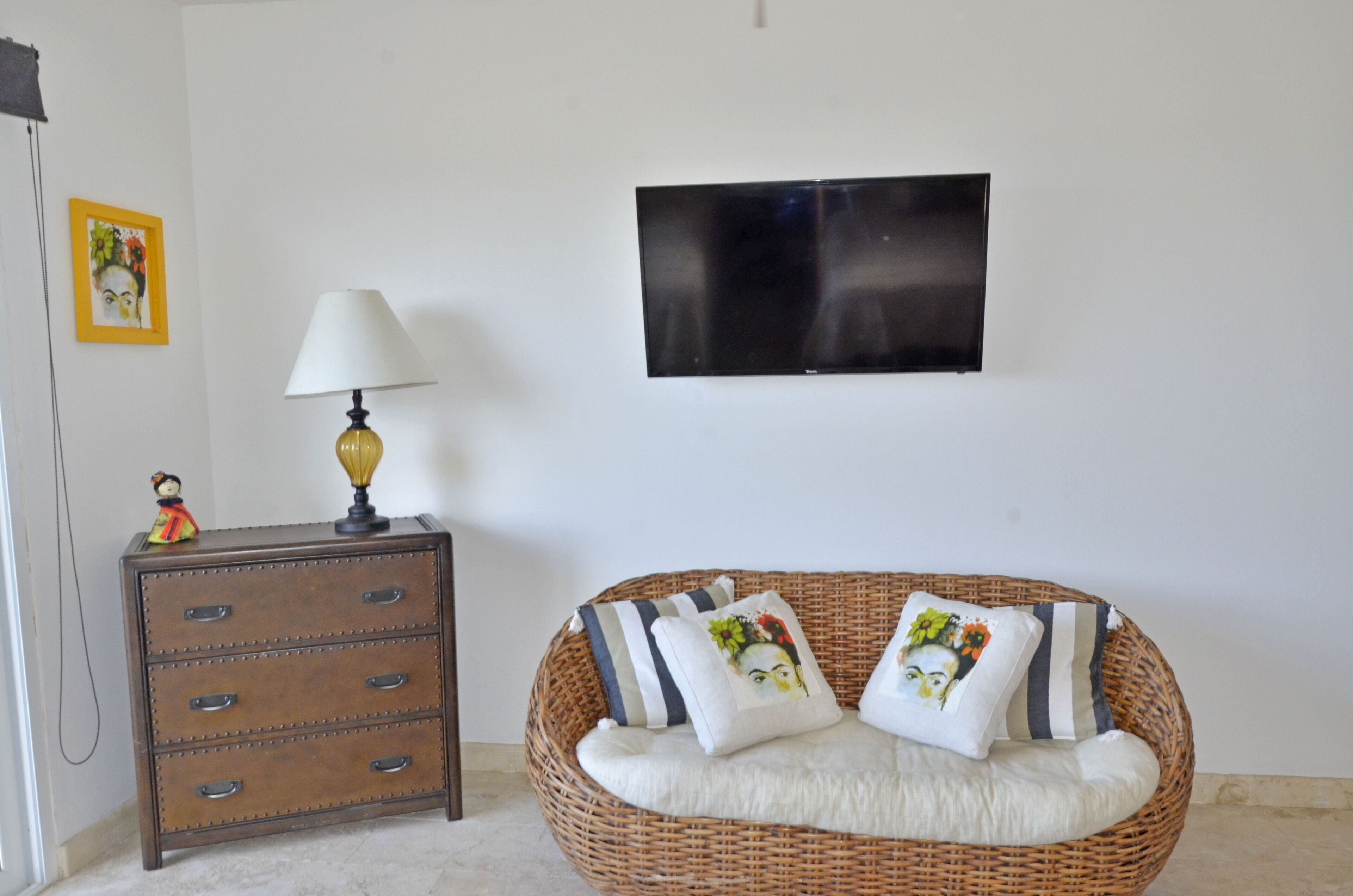 Una habitación con un sofá de mimbre, un aparador de madera con cajones, un televisor montado en la pared y una mesita con una lámpara y una figurita.