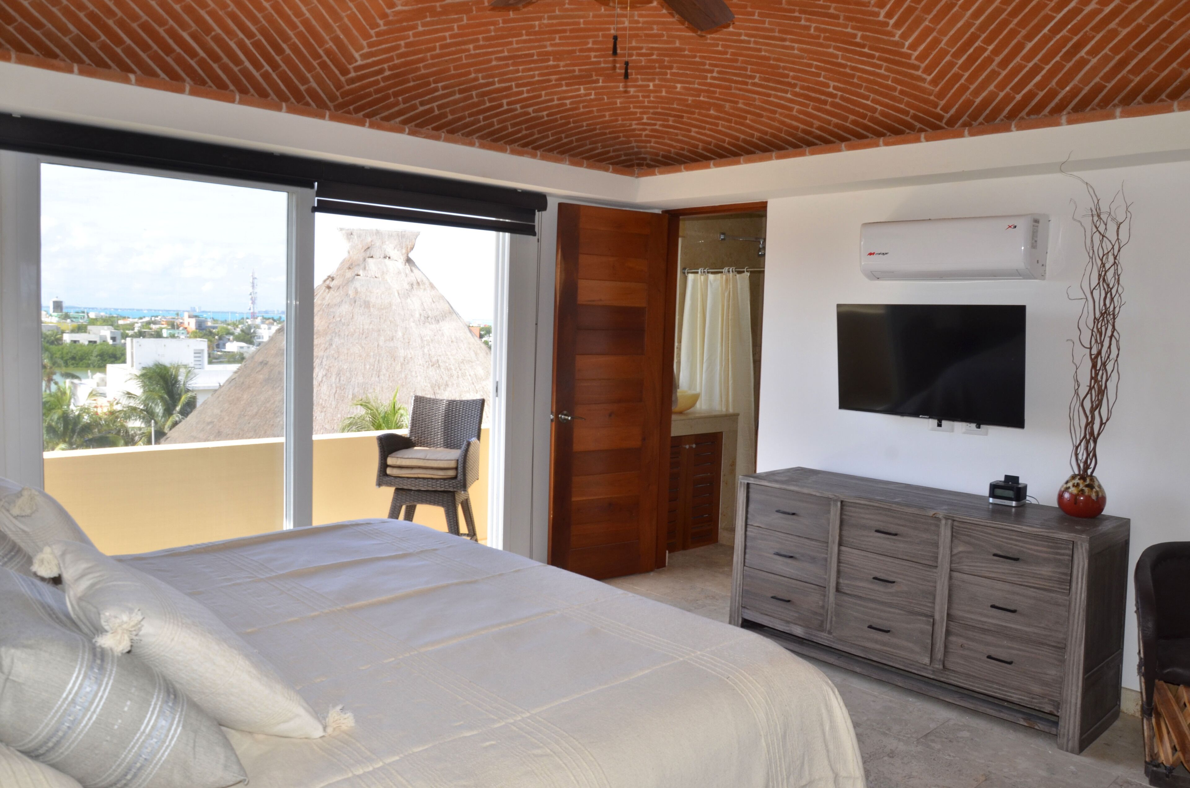 Un dormitorio con una cama grande, televisor, cómoda de madera y puerta corrediza de vidrio que da a un balcón con vista a la ciudad.