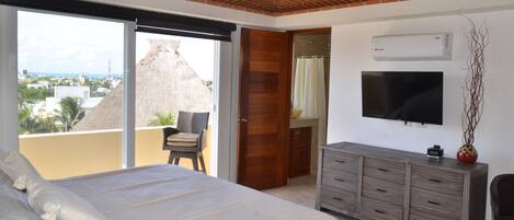 Un dormitorio con una cama grande, televisor, cómoda de madera y puerta corrediza de vidrio que da a un balcón con vista a la ciudad.
