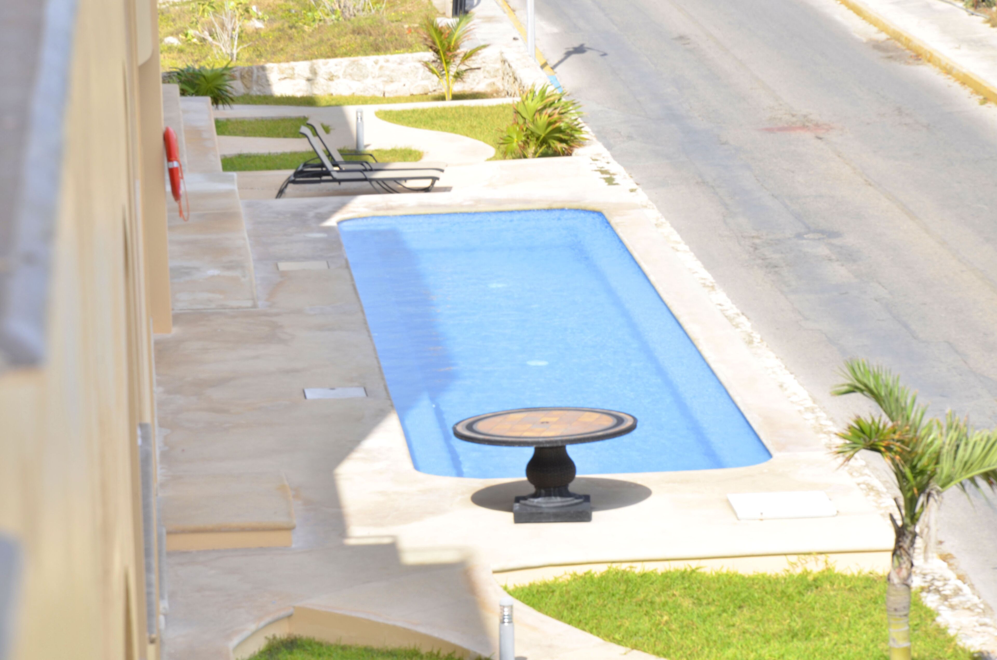 A rectangular swimming pool with a small table in the center, surrounded by a paved area with a few chairs and small palm trees.