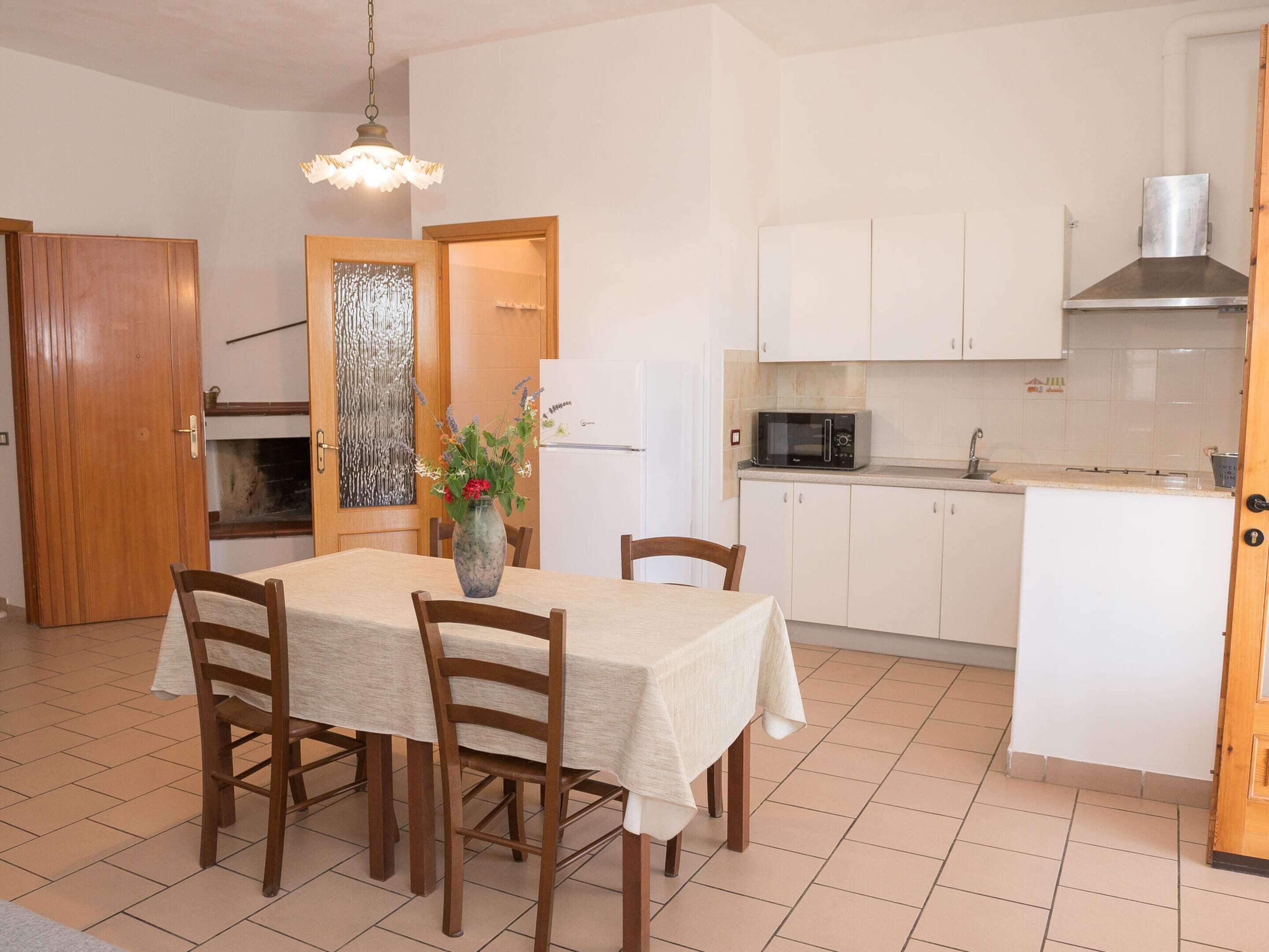 Cucina compatta con mobili bianchi, un forno a microonde e un lavandino. Si nota un tavolo da pranzo con sedie e un vaso con fiori.
