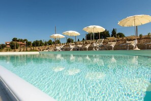 Piscina cristalina con sillas de descanso y sombrillas, rodeada por una valla y árboles.