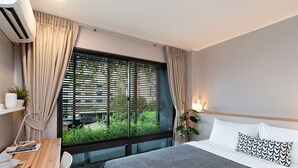 A modern bedroom with a large window, a bed with patterned pillows, a bedside table with a lamp, and a potted plant. - GROOVE AT SIAM (Bangkok)