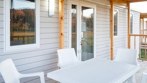 A covered patio area with a white table and chairs, wooden beams, and windows overlooking a natural landscape.
