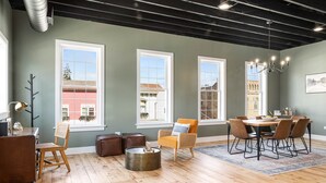 A spacious living area with a wooden floor, a dining table with chairs, a sofa, a coffee table, and large windows offering views of buildings outside.