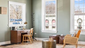 A room with a desk, chair, and a wicker chair. There are windows with views of buildings and trees.