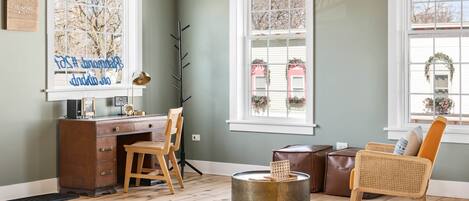 Una habitación con escritorio, silla y sillón de mimbre. Se ven edificios y árboles a través de las ventanas.
