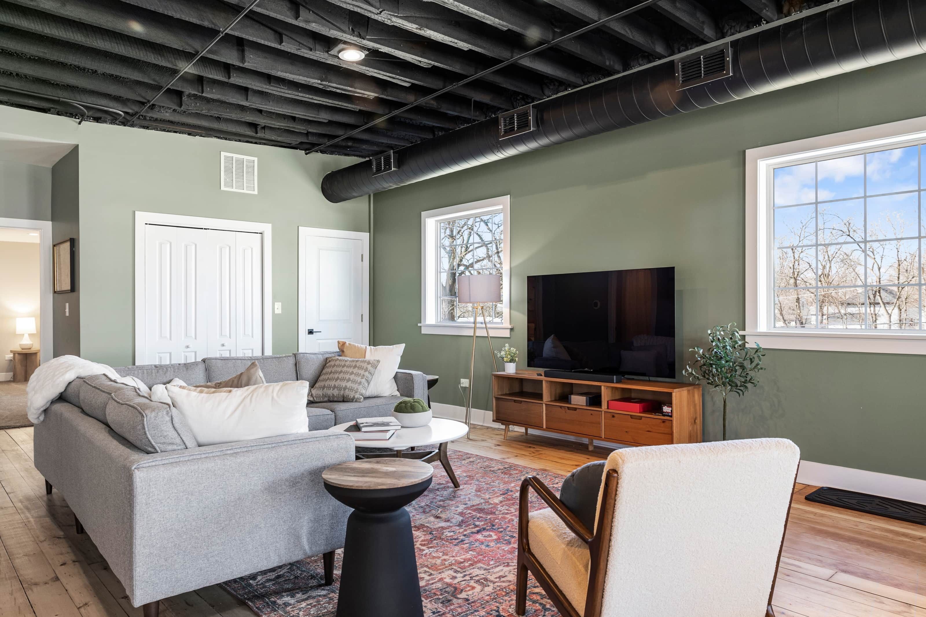 A living room with a grey sofa, a coffee table, and a TV. There are windows, a door, and a plant.