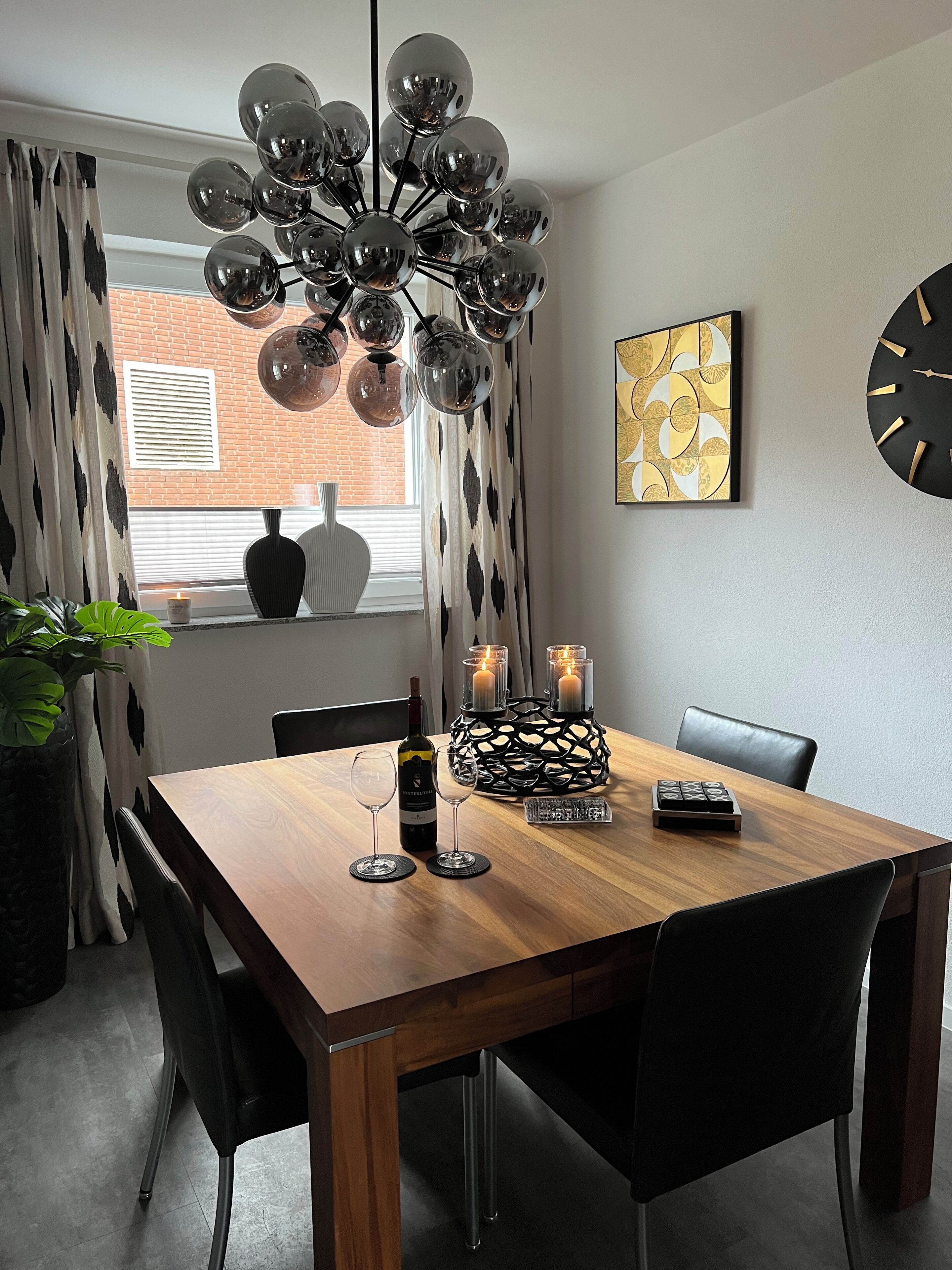 A dining area with a wooden table, black chairs, a modern chandelier, candles, and a decorative vase.