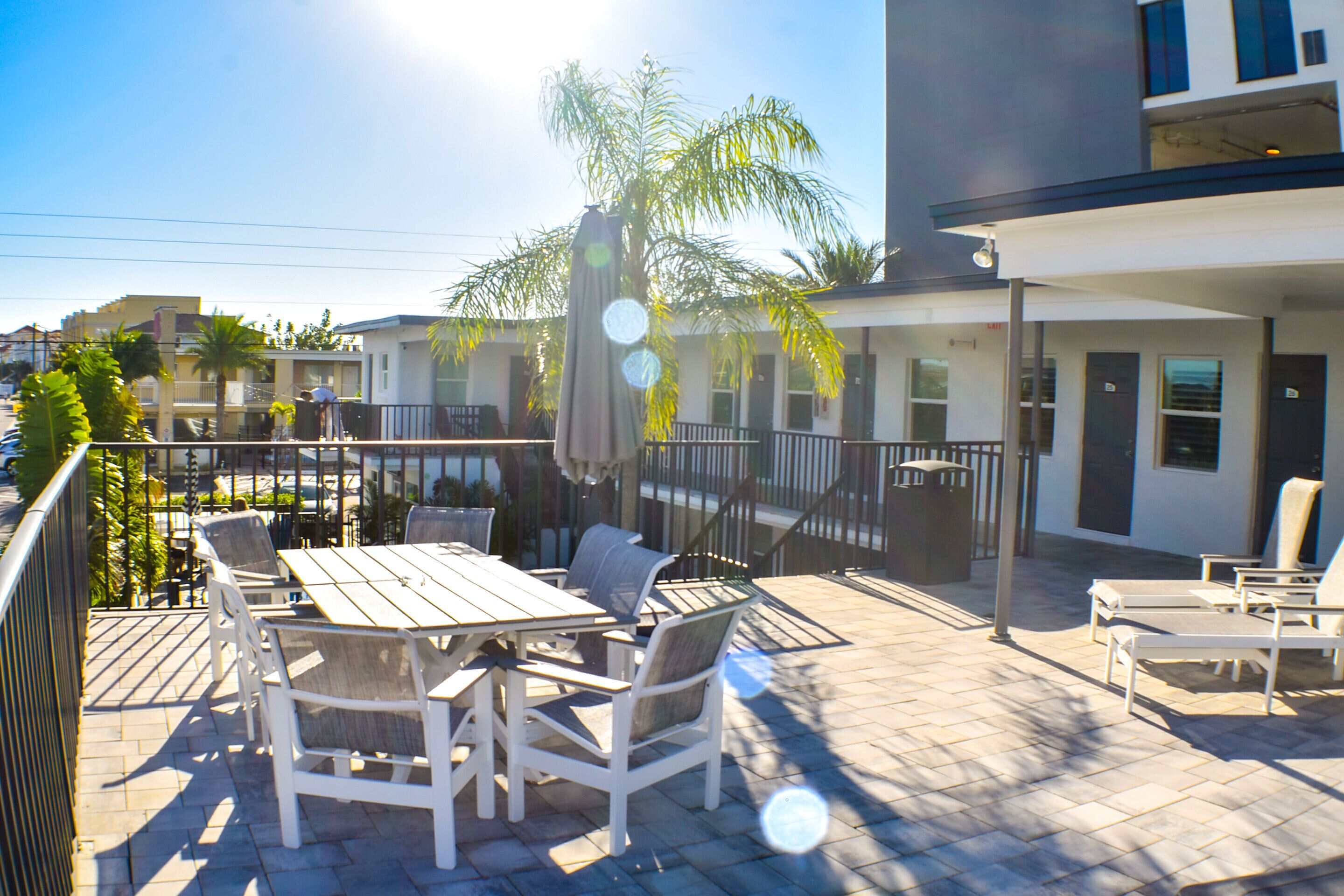 An outdoor patio with a wooden table and chairs, surrounded by palm trees and a modern building in the background.