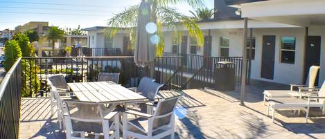 An outdoor patio with a wooden table and chairs, surrounded by palm trees and a modern building in the background.