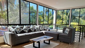 A modern living room with a grey sectional sofa, a wooden coffee table, and a grey armchair. Large windows offer a view of palm trees and a pool. - Private Beach side Home - Playa San Diego (La Libertad)