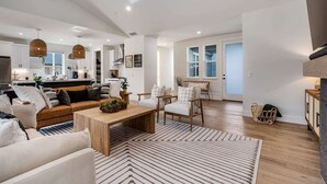 A spacious living area with a sofa, coffee table, and chairs, a kitchen in the background, and a large window.