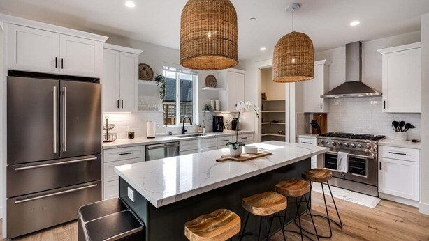 A modern kitchen with white cabinetry, stainless steel appliances, a large island with stools, and wicker pendant lighting.
