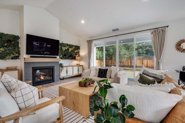 A modern living room with a fireplace, a flat-screen TV mounted above it, and a large window offering a view of the outdoors. - Boho Betty (Windsor)
