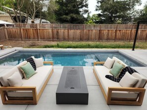 A poolside area with two sofas and a table, surrounded by a wooden fence and trees. - Boho Betty (Windsor)