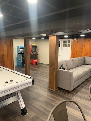 A pool table, a sofa, a red arcade machine, and a door in a room with wooden walls and a ceiling.