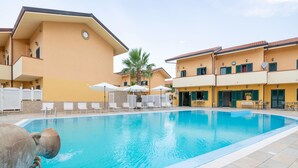 Una piscina con sillones de playa y sombrillas, rodeada de edificios amarillos con balcones.