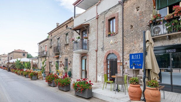 Una hilera de edificios de piedra tradicionales con balcones, mesas al aire libre y macetas con plantas.