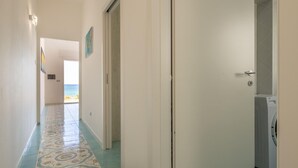 Ein Flur mit gemusterten Bodenfliesen, eine Tür zum Waschbereich und Blick auf das Meer durch eine offene Tür am Ende.