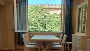 A dining area with a table and chairs, a window with curtains, and a view of greenery and buildings outside. - Holiday Home in Reggio Calabria Near Beach (Reggio Calabria)