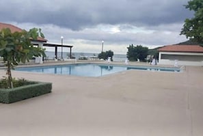 Una piscina con vista al mar, rodeada por una zona cubierta con asientos y un pequeño jardín.