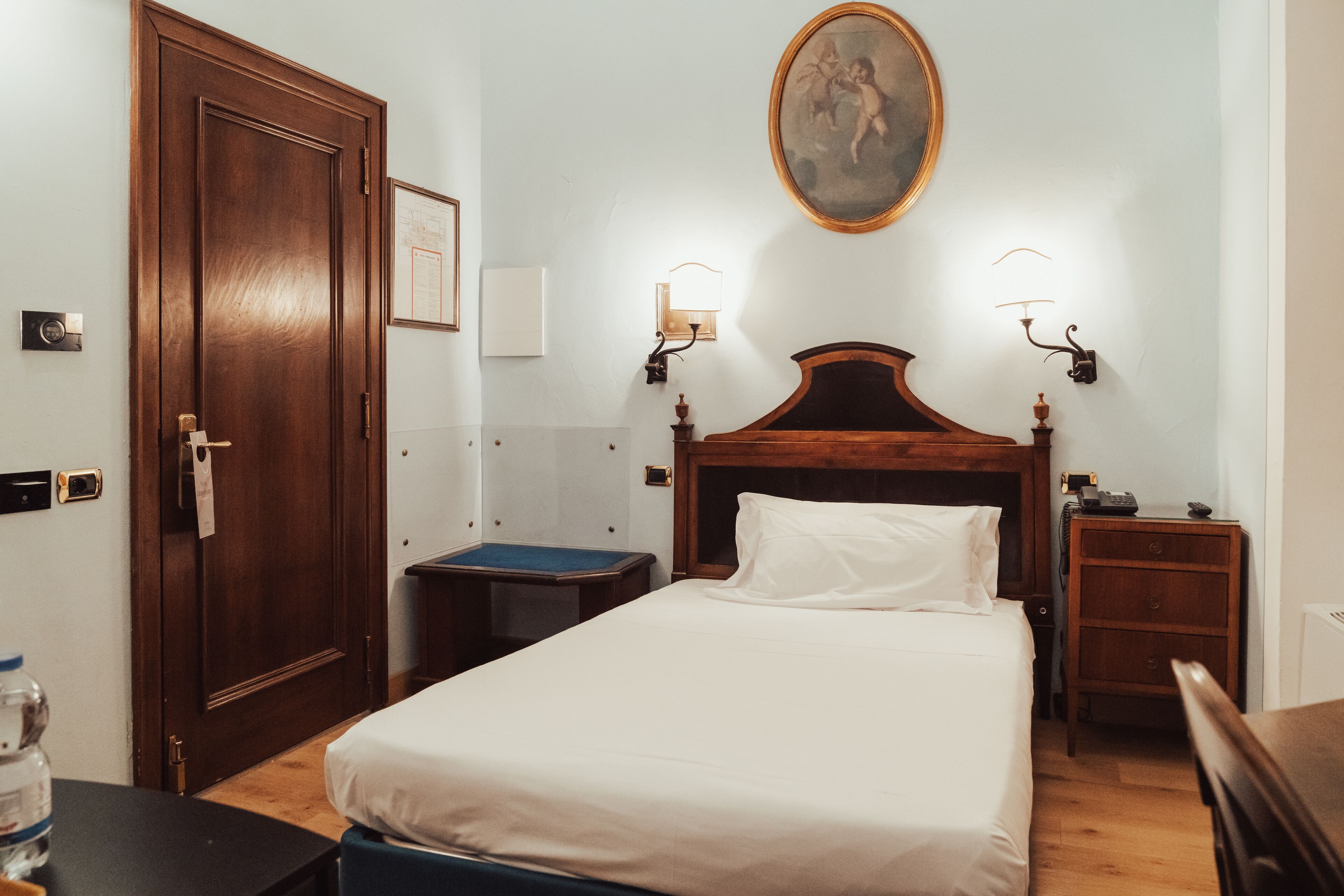 A hotel room with a wooden bed, a nightstand, a desk, a chair, a water bottle, and a door.