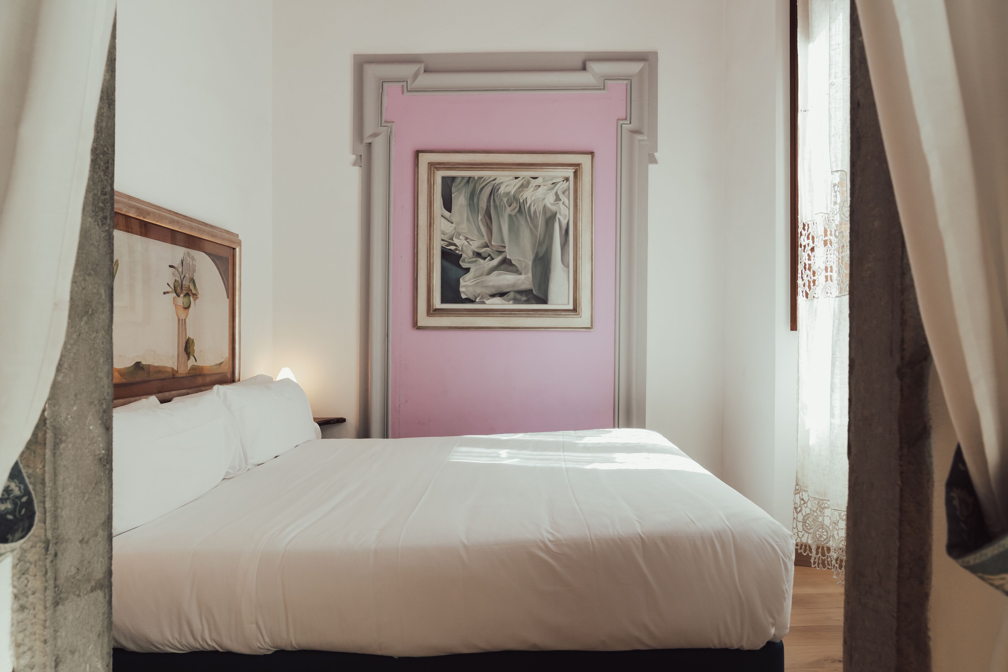 Ein ordentlich bezogenes Bett in einem Zimmer, dessen Wand in Rosa gehalten ist, ein gerahmtes Kunstwerk über dem Bett und ein Fenster mit transparenten Vorhängen.