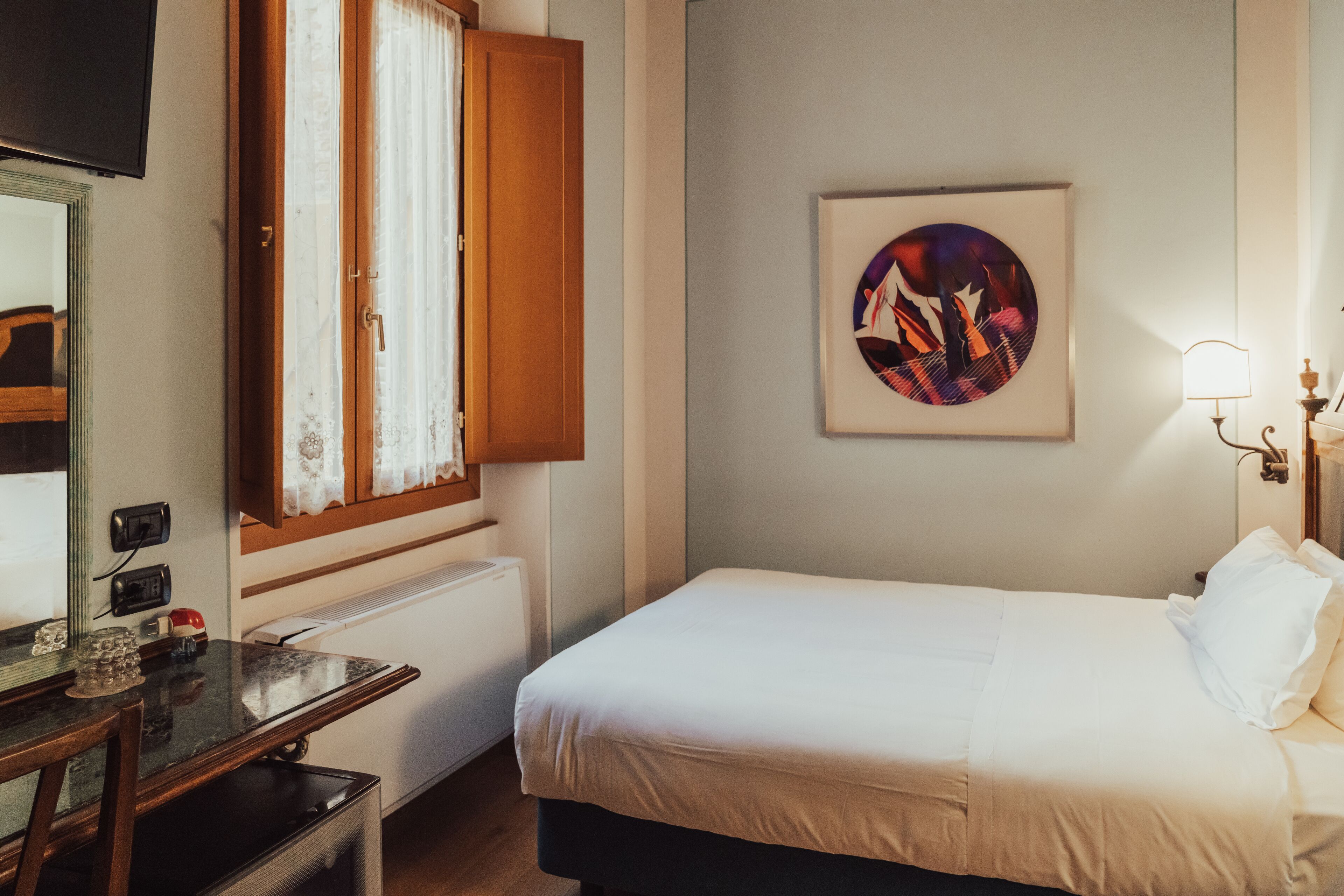 A hotel room with a bed, a desk with a chair, a window with curtains, a wall-mounted picture, and a bedside lamp.