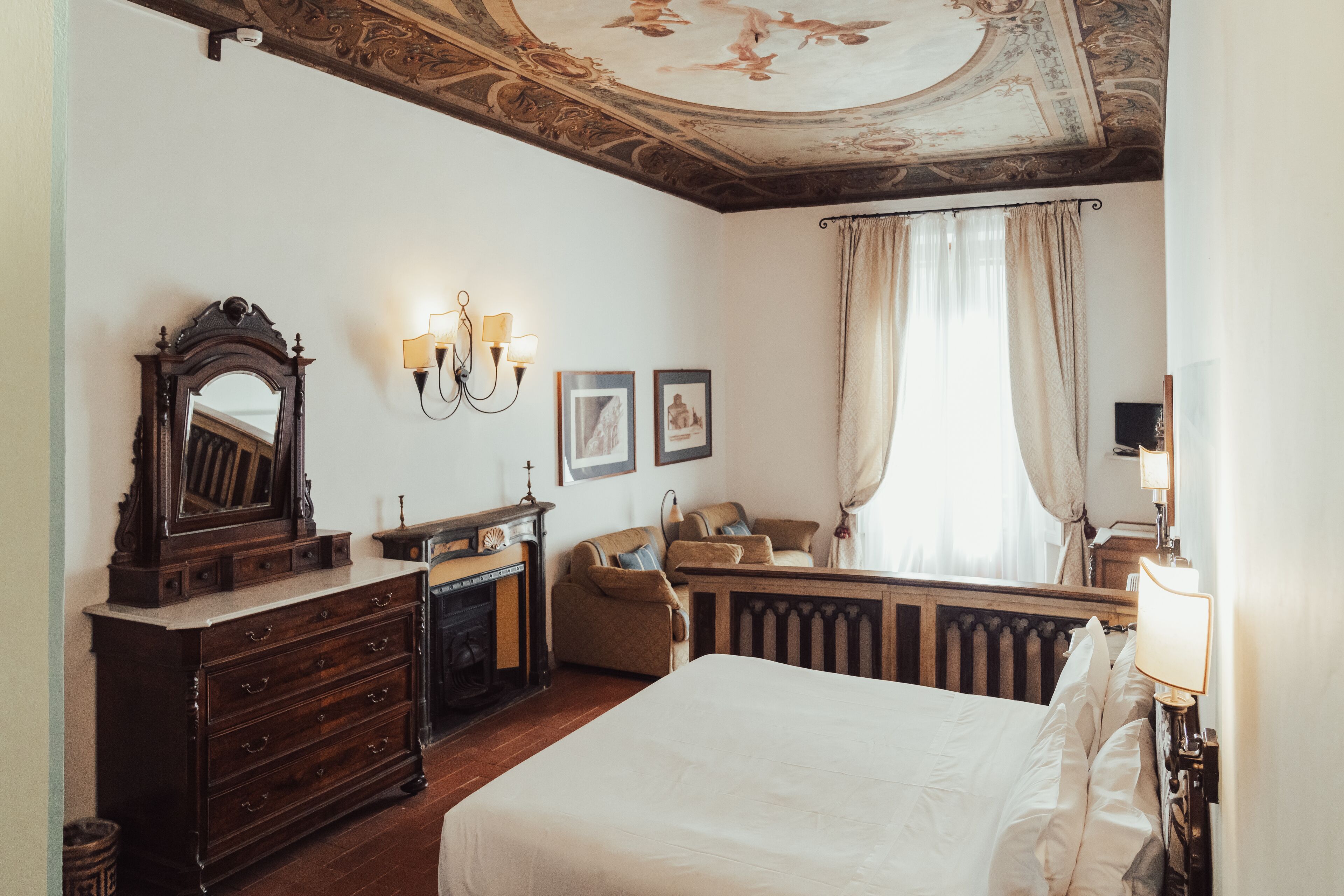 A bedroom with a large bed, a dresser, a mirror, a fireplace, and a chandelier.
