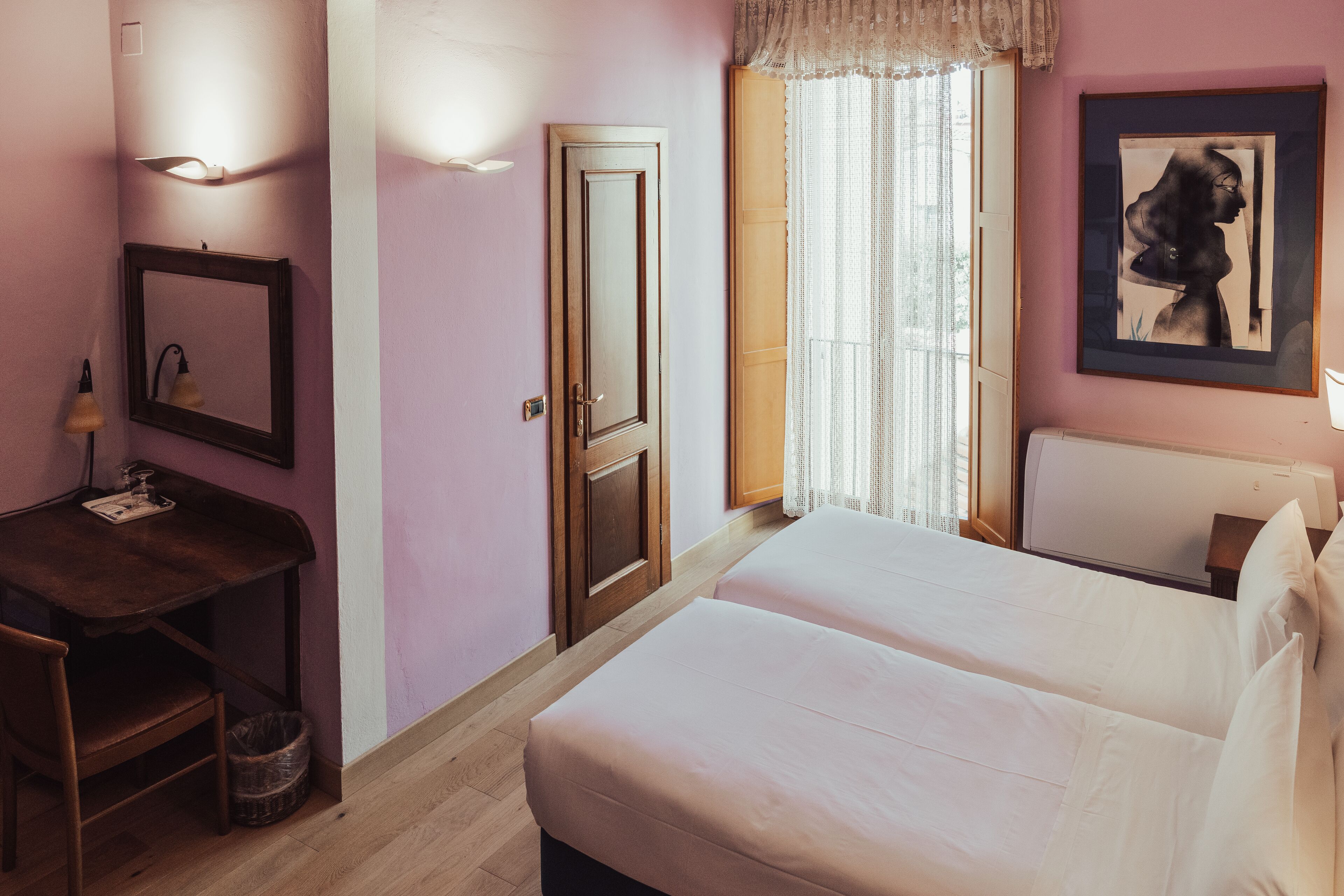 Ein Hotelzimmer mit einem Bett, einem Schreibtisch, einem Stuhl, einem Mülleimer, einer Tür, einem Fenster mit Vorhängen und einem gerahmten Kunstwerk an der Wand.