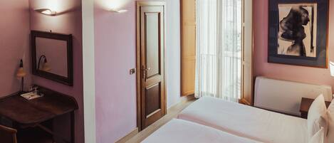 A hotel room with a bed, a desk, a chair, a trash bin, a door, a window with curtains, and a framed artwork on the wall.