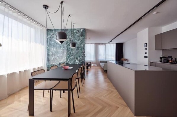 Une cuisine moderne avec un pare-feu en marbre, un coin repas avec une table et des chaises noires, et une grande fenêtre agrémentée de rideaux légers.