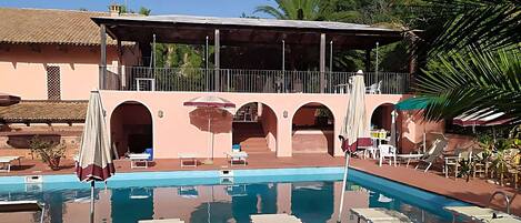A pool area with lounge chairs and umbrellas, a covered seating area, and a building with arches.