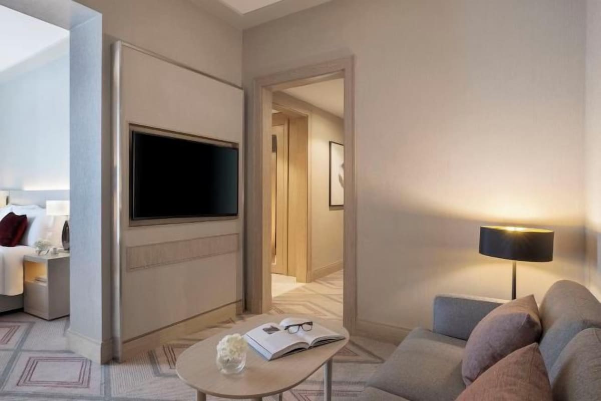 A hotel room with a flat-screen TV, a sofa, a small table with a book and glasses, and a lamp.