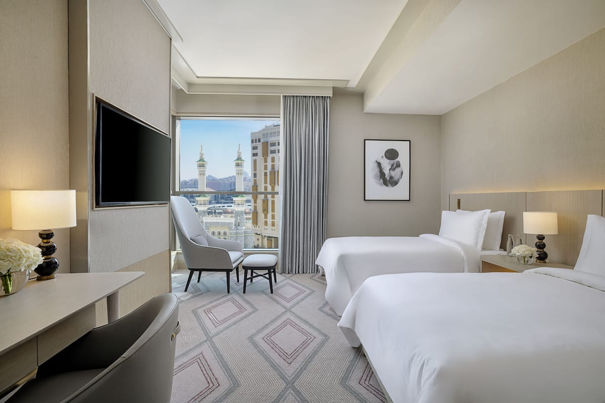A hotel room with two beds, a desk, a chair, a TV, and a large window offering a city view.