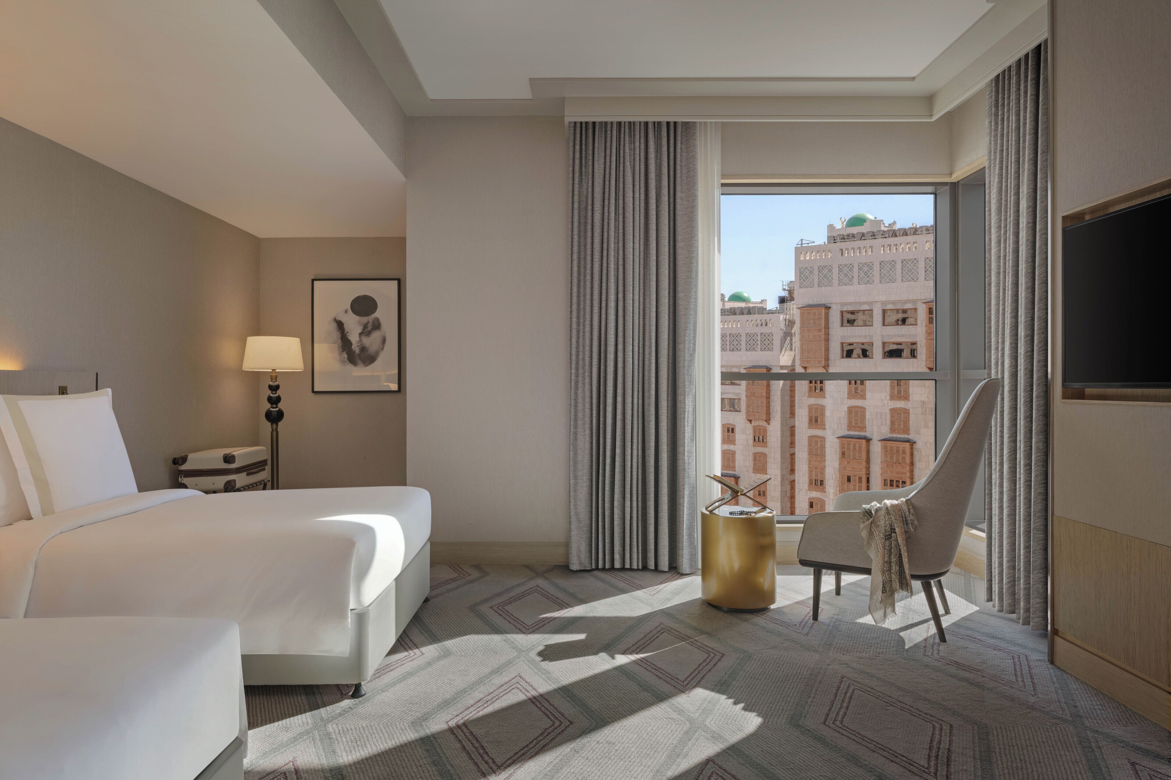 Hotelkamer met een bed, stoel, lamp, schilderij aan de muur en uitzicht op gebouwen door het raam.