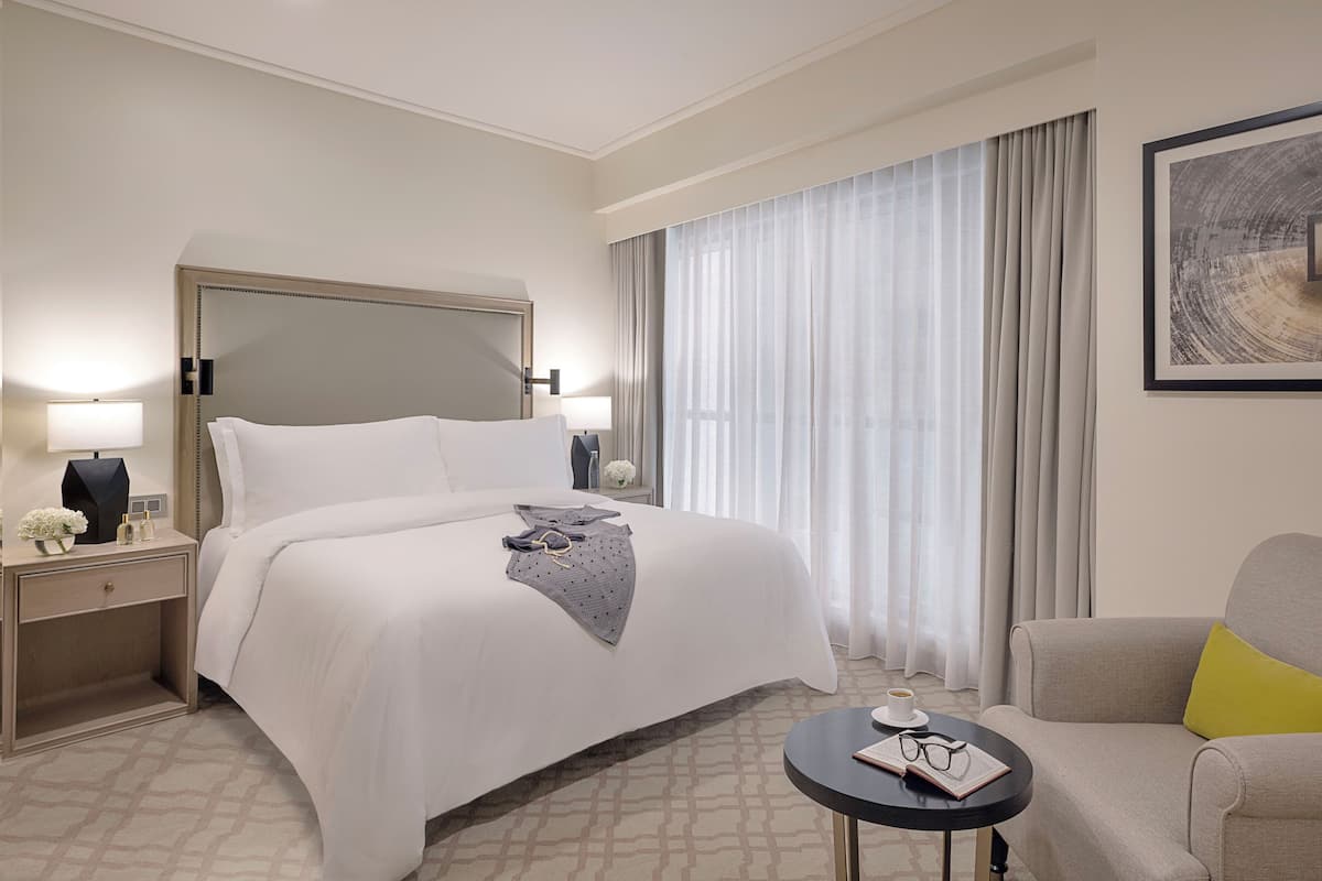 A hotel room with a large bed, a bedside table with a lamp, a small table with a book and glasses, a sofa with a yellow pillow, and a large window with curtains.