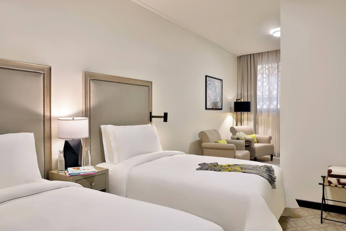 A hotel room with two beds, a sitting area with a chair, a TV, and a large mirror.
