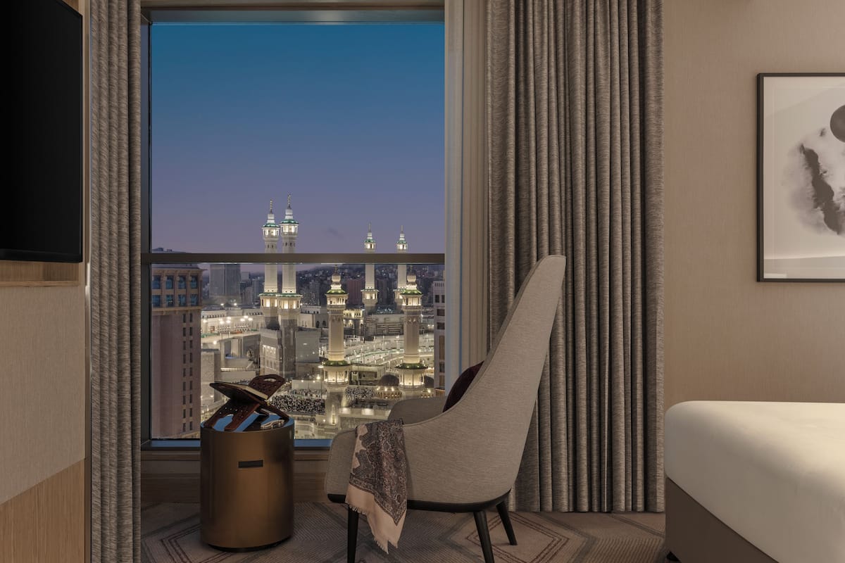 A hotel room with a large window offering a view of a cityscape at dusk, a comfortable armchair, a bedside table with a lamp, and a neatly made bed.