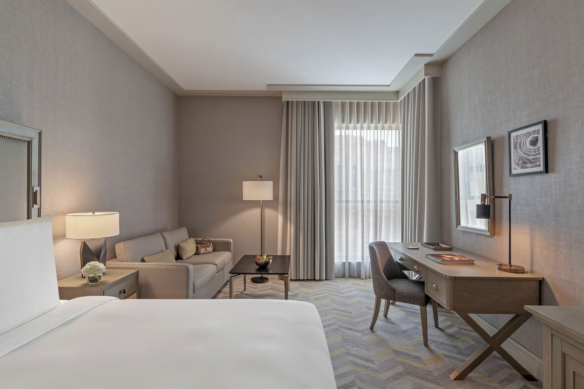 A hotel room with a bed, a desk, a chair, a nightstand, a lamp, a mirror, and a window with curtains.