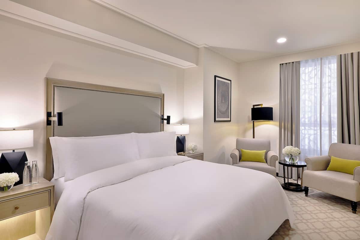 A hotel room with a large bed, a sitting area with a chair and a small table, a nightstand with a lamp, and a window with curtains.